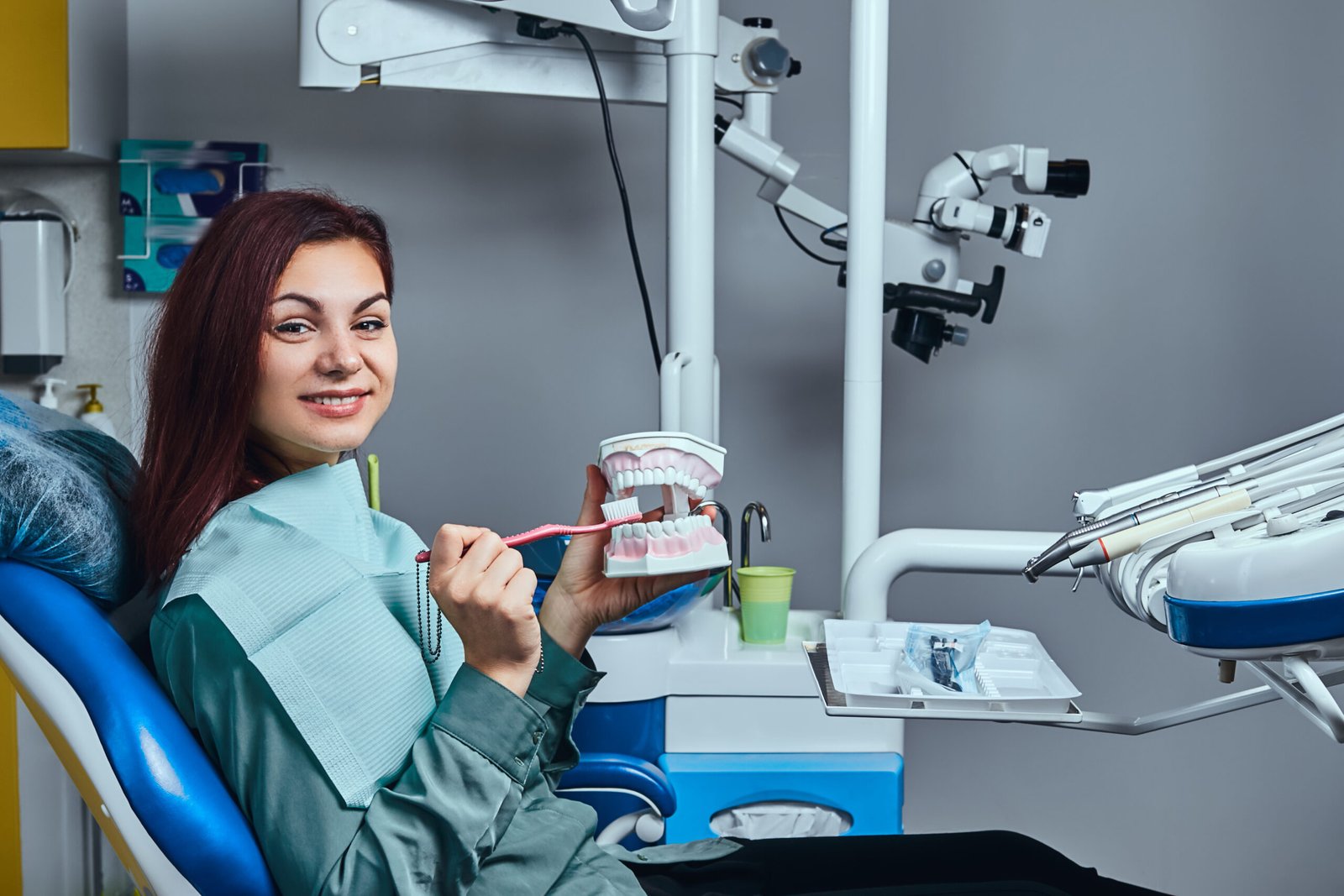 Happy young redhead woman sitting in a dentist chair and holding a toothbrush and denture in a dentist office.